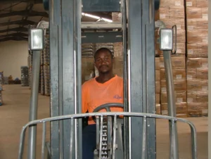 A forklift operator at the wheel of a forklift inside a warehouse stacked with palletized boxes