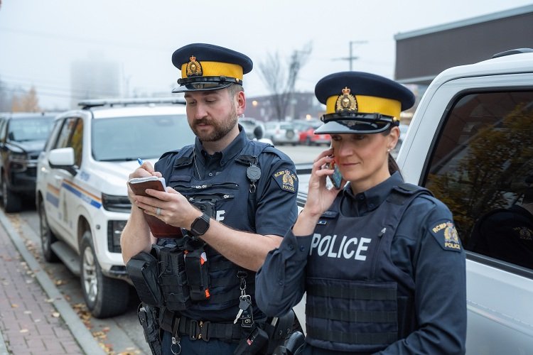 Two RCMP officers in navy-blue uniform shirts beside police vehicles, one writing notes and one on the phone. Photo: BC RCMP
